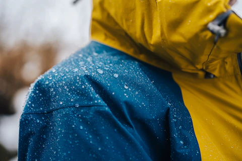 Close-up of a waterproof jacket with yellow and blue panels, covered in water droplets, indicating it is being worn in wet conditions